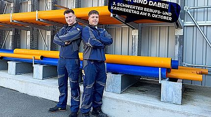 Zwei Auszubildende der Stadtwerke Schwerte in Arbeitskleidung stehen Rücken an Rücken vor einem Regal mit gelben und blauen Rohren. Daneben eine Sprechblase mit dem Hinweis: ‚Wir sind dabei! 3. Schwerter Berufs- und Karrieretag am 9. Oktober‘.
