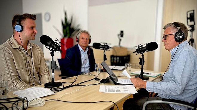 Drei Männer sitzen mit Kopfhörern und Mikrofonen in einem Studio und sprechen miteinander. Vor ihnen liegen Notizen und ein Laptop – es handelt sich um eine Podcast-Aufnahme.