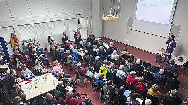 Eine Präsentation in einem Seminarraum mit vielen Teilnehmern, die auf Stühlen sitzen und einem Redner zuhören, der an einem Pult steht und eine Präsentationsfolie zeigt.