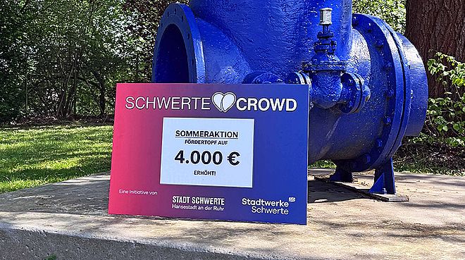 Ein großes, blaues Wasserrohr steht auf einem Betonpodest vor einer Baumreihe, davor befindet sich ein Schild mit der Aufschrift "Schwerte Crowd Sommeraktion Fördertopf auf 4.000 € erhöht".