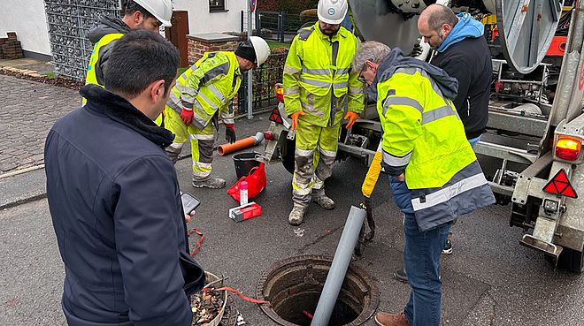 Mehrere Menschen stehen um einen geöffneten Kanaldeckel in der Elsetalstraße. Ein lange Rohr schaut aus dem Kanaldeckel heraus. Hier wird die neue Technik zur Sanierung von Abwasserdruckleitungen von der Stadtentwässerung Schwerte durchgeführt.