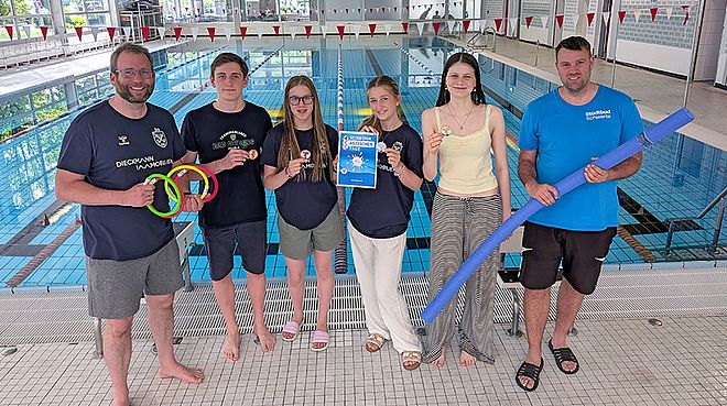 Eine Gruppe von sechs Personen steht im Stadtbad Schwerte und hält verschiedene Schwimmutensilien sowie Schwimmabzeichen hoch. Symboldbild für den Schwimmabzeichentag 2026 im Stadtbad Schwerte am 14. Juni 2026.