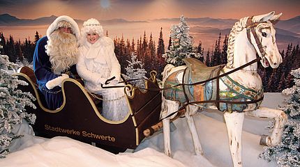 Ein Weihnachtsmann im blauen Kostüm sitzt mit einem Engelchen in einem Schlitten, davor ist ein Pferd aus Holz. Der Schlitten steht in einer künstlichen Winterlandschaft.