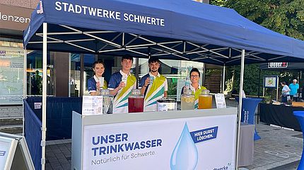 Infostand der Stadtwerke Schwerte beim Pannekaukenfest: Vier junge Mitarbeiterinnen und Mitarbeiter schenken verschiedene Getränke aus großen Gläsern aus. Am Stand mit blauem Pavillon wird das Motto ‚Unser Trinkwasser – Natürlich für Schwerte‘ präsentiert.