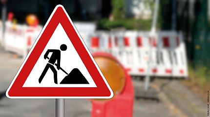 Hier entstehen kurzfristig Baustellen. Im Vordergrund sieht man das Verkehrsschild für Baustellen. Im Hintergrund ist unscharf eine Baustelle zu erkennen.