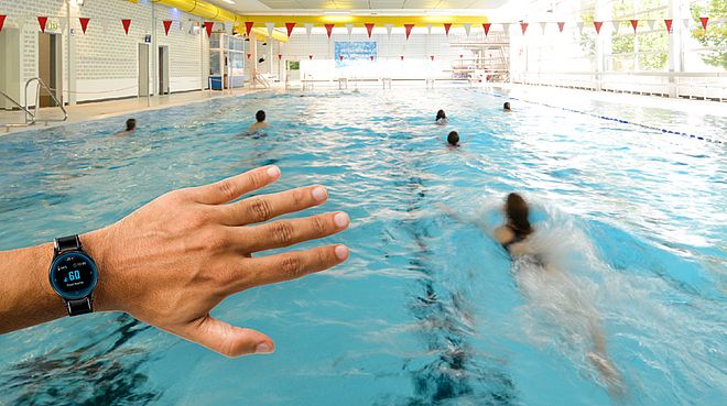 Auf dem Bild sieht man im Hintergrund das Schwimmerbecken vom Stadtbad Schwerte. Im Vordergrund steht eine Hand mit einer speziellen Armbanduhr, um die Informationen der KI zu empfangen. 