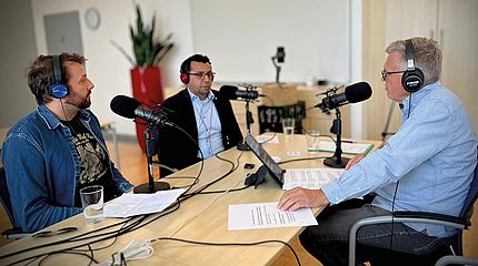 Drei Männer mit Kopfhörern und Mikrofonen sitzen an einem Tisch und nehmen eine Podcastfolge auf. Vor ihnen liegen Notizen und Gläser Wasser.