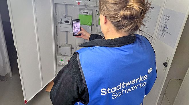 Auf dem Bild steht eine Frau mit einer Weste der Stadtwerke Schwerte vor einem Zählerschrank und fotografiert mit ihrem Handy den Stromzähler.