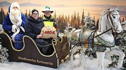 Drei Personen sitzen in einem Schlitten der Stadtwerke Schwerte vor einer winterlichen Landschaft mit vielen Bäumen und Schnee. Der Schlitten wird von einem Holzpferd gezogen. 