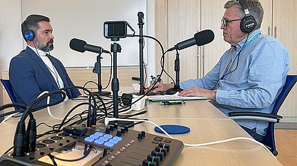 Zwei Personen sitzen sich an einem Tisch gegenüber. Auf dem Tisch steht ein kleines Mischpult, Mikrofone und eine Kamera. Die Szene zeigt die Aufnahme eines Podcasts. 