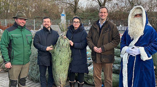 Vier Personen und ein Mann in einem blauen Weihnachtsmannkostüm stehen vor eingepackten Tannenbäumen. 
