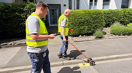 Zwei Männer mit gelben Westen schieben eine Teppichsonde über den Asphalt.
