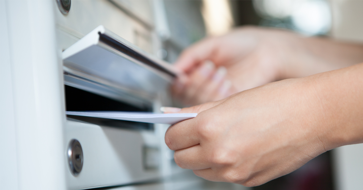 Eine Hand öffnet einen Briefkasten und wirft einen Brief ein.