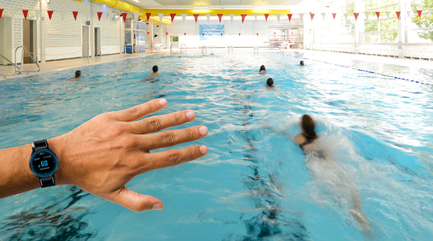 Auf dem Bild sieht man im Hintergrund das Schwimmerbecken vom Stadtbad Schwerte. Im Vordergrund steht eine Hand mit einer speziellen Armbanduhr, um die Informationen der KI zu empfangen. 