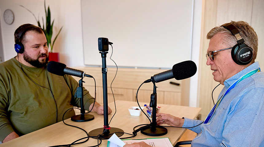 Zwei Männer sitzen an einem Tisch und nehmen mit Mikrofonen und Kopfhörern eine Podcast-Folge auf; zwischen ihnen steht eine kleine Kamera, während einer spricht und der andere mit Notizen zuhört.