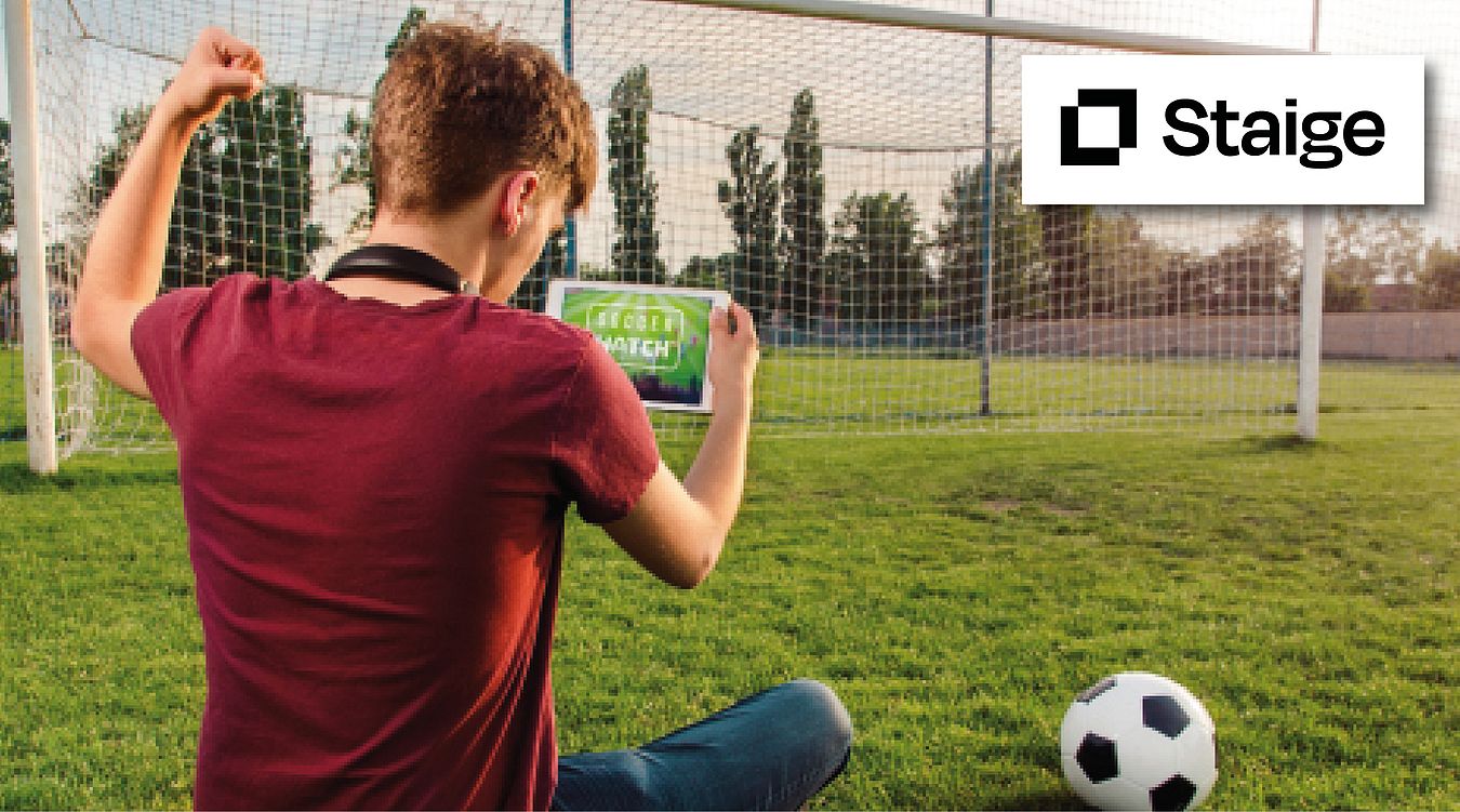 Ein Junge sitzt auf dem Fußballplatz vor dem Tor und schaut auf sein Handy, wo eine Übertragung läuft. Symbolbild für die Übertragungen im Soccerwatch TV