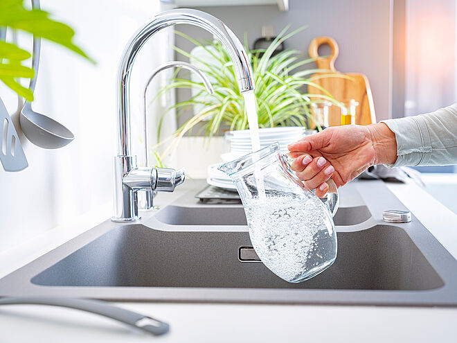 Eine Hand hält eine Glaskaraffe unter den laufenden Wasserhahn als Symbolbild für unser Wasser in Schwerte.