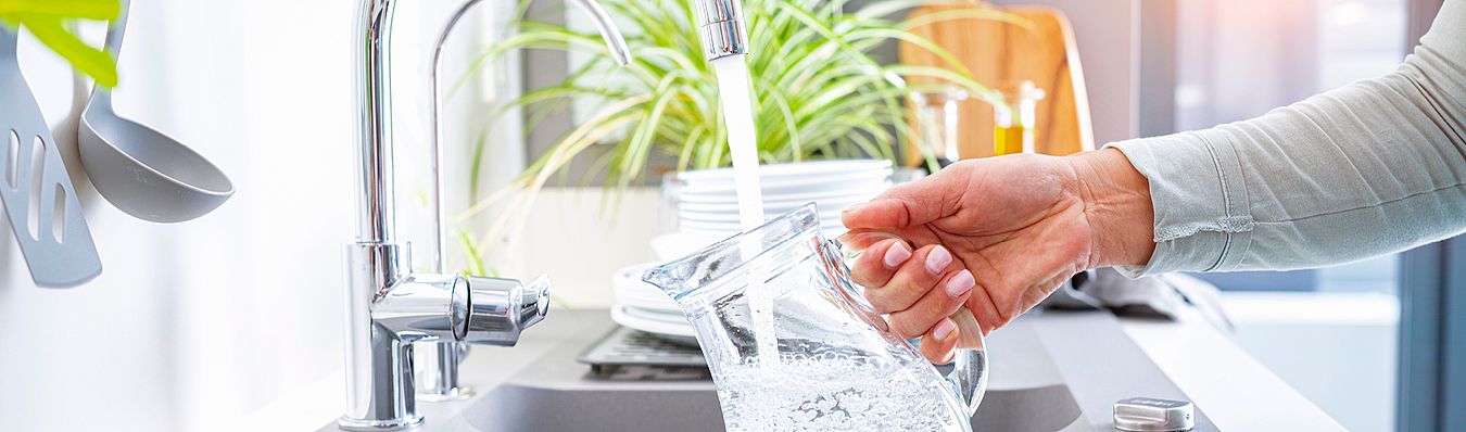 Eine Hand hält eine Glaskaraffe unter den laufenden Wasserhahn als Symbolbild für unser Wasser in Schwerte.