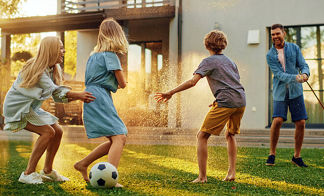 Drei Kinder spielen im Garten mit einem Fussball. Ein Mann hält einen Gartenschlauch. Symbolbild für Eigentümer in Schwerte