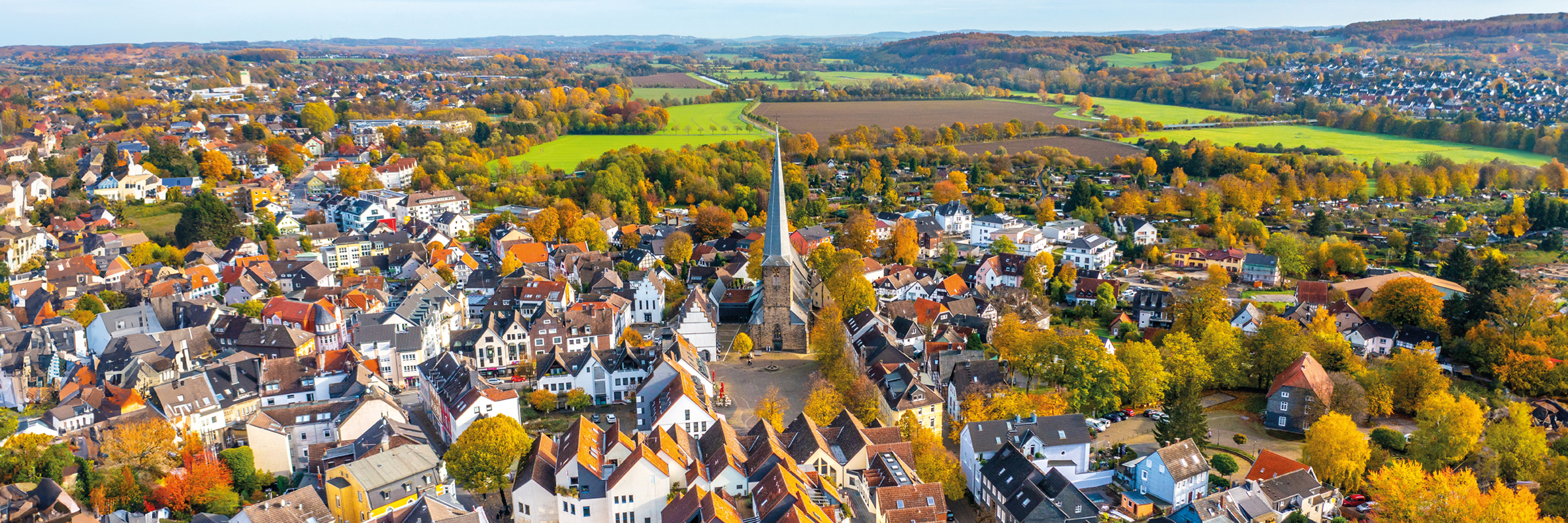 Luftaufnahme der Stadt Schwerte mit der Sankt-Viktor-Kirche im Zentrum, umgeben von Häusern und herbstlich gefärbten Bäumen, im Hintergrund Felder und Hügel